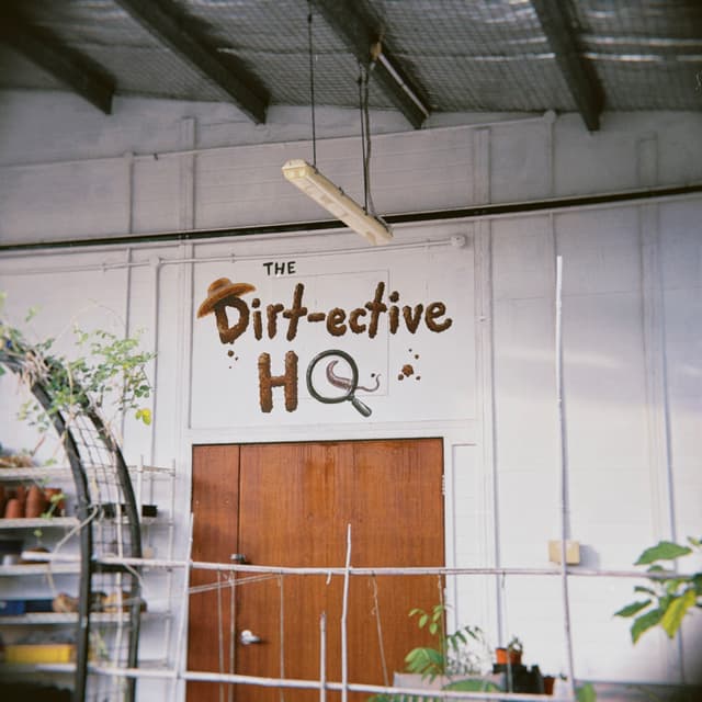 A white wall with wood double doors has the words 'The Dirt-ective HQ' painted on it A white light fixture hangs from the slanted ceiling above Green plants with a white railing are partially visible in front of the doors