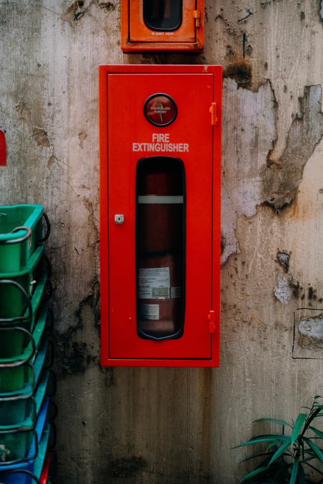 A red metal box mounted on a concrete wall holds a fire extinguisher and has a glass window, with faded white text that reads FIRE EXTINGUISHER