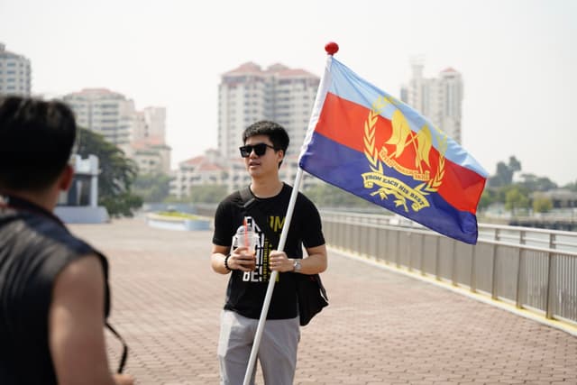 A young man stands outdoors holding a pole with a flag, appearing to have a conversation with another individual who is out of focus in the foreground. Urban buildings can be seen in the background