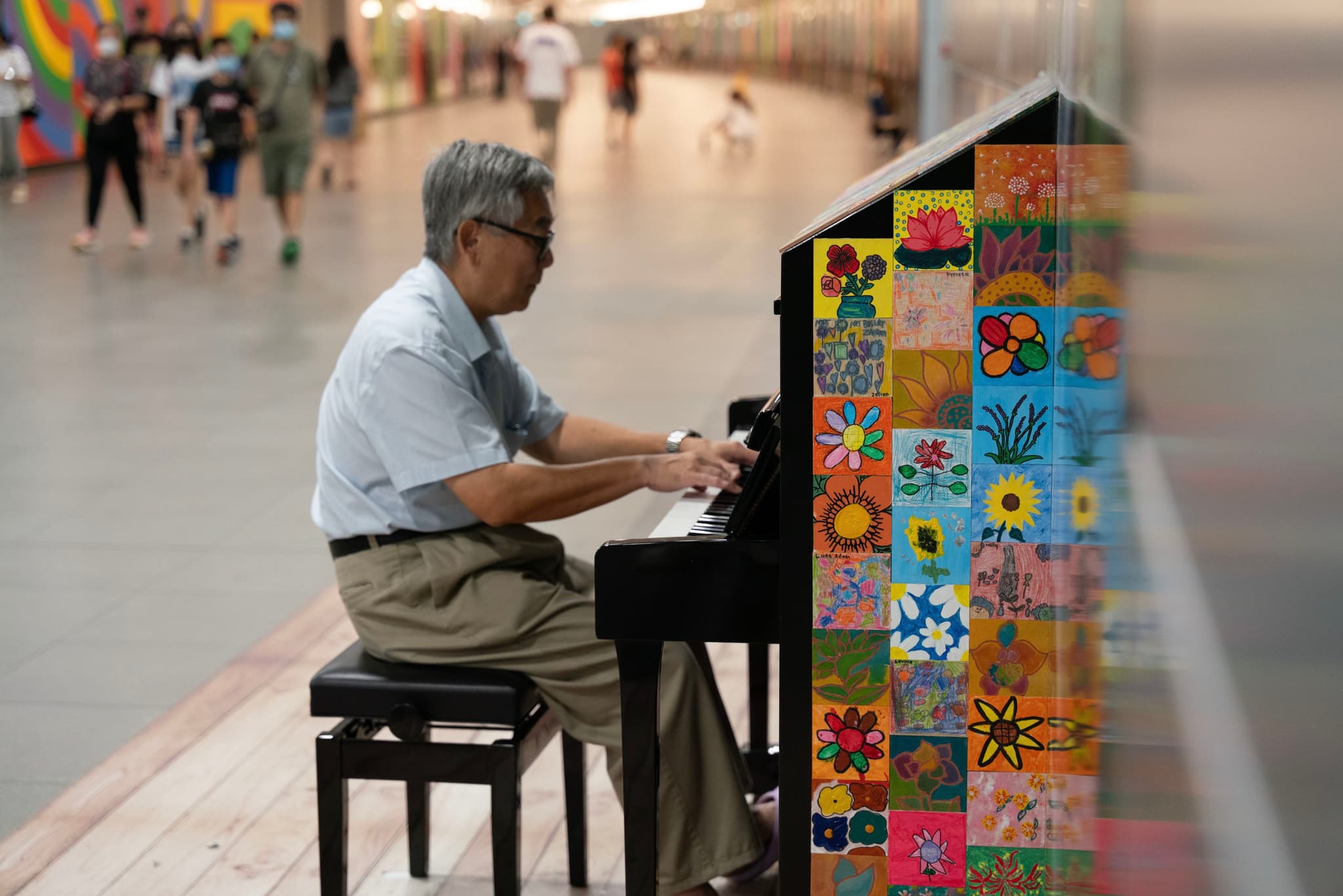 An elderly man is playing a colorfully decorated upright piano in a public space, while blurred figures walk by in the background