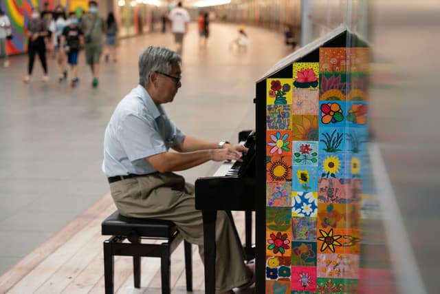 An elderly man is playing a colorfully decorated upright piano in a public space, while blurred figures walk by in the background