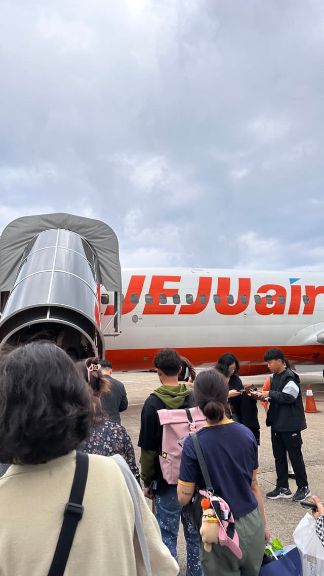 A group of people stand on the tarmac facing an airplane with a red and white fuselage that reads JEJUair. A covered jetway is to the left, and the sky is cloudy