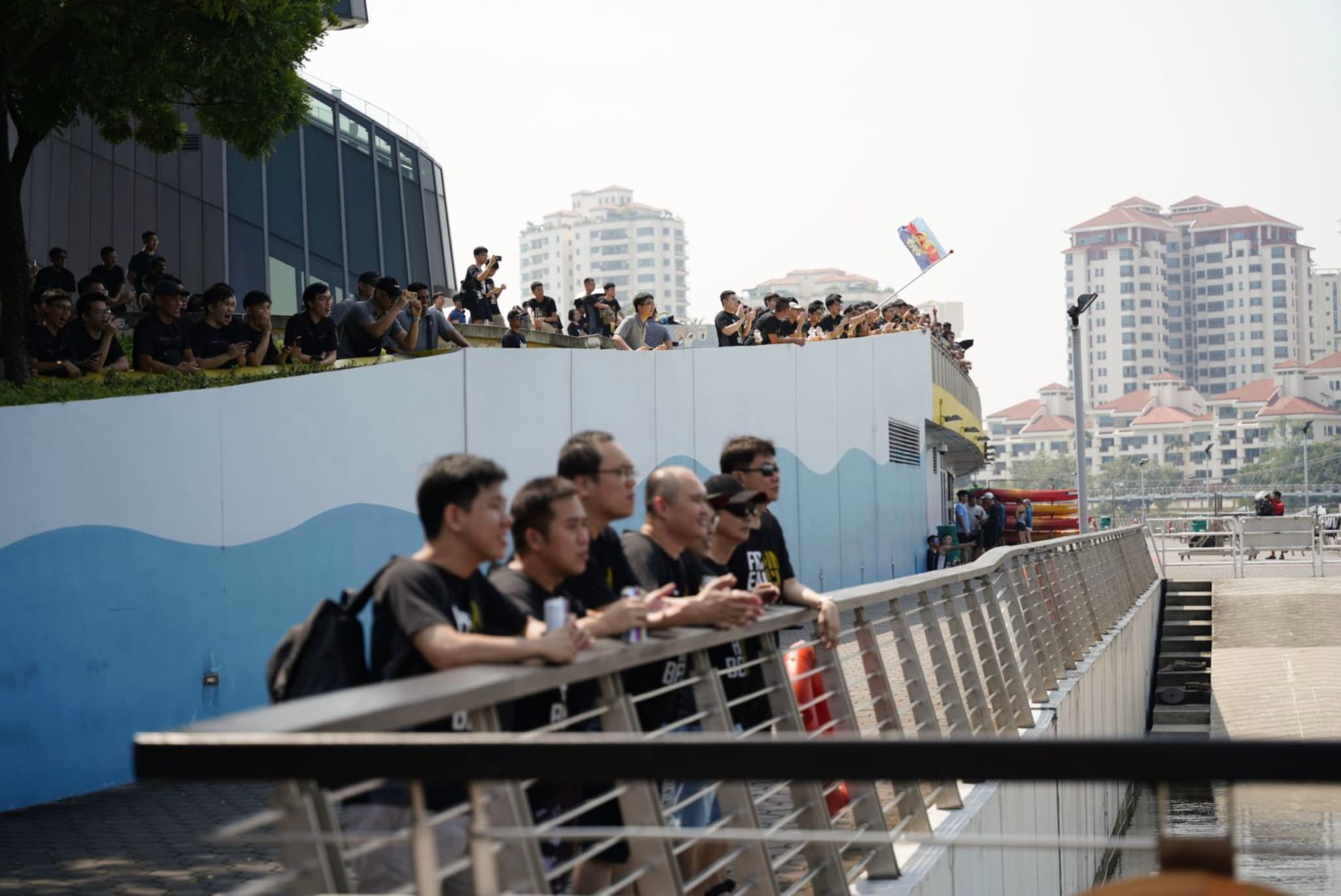 A group of spectators is gathered behind a railing, likely watching an event, with more people visible on an elevated grassy area in the background, against an urban backdrop with apartment buildings