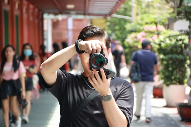 A person with a camera covering their face stands in the foreground, taking a photo, with a blurred background of people walking on a street