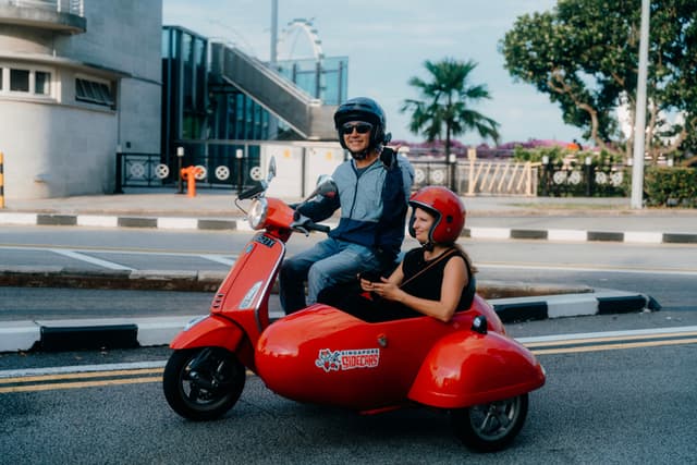A man and a woman ride a red motor scooter with a sidecar down a city street, both wearing helmets for safety