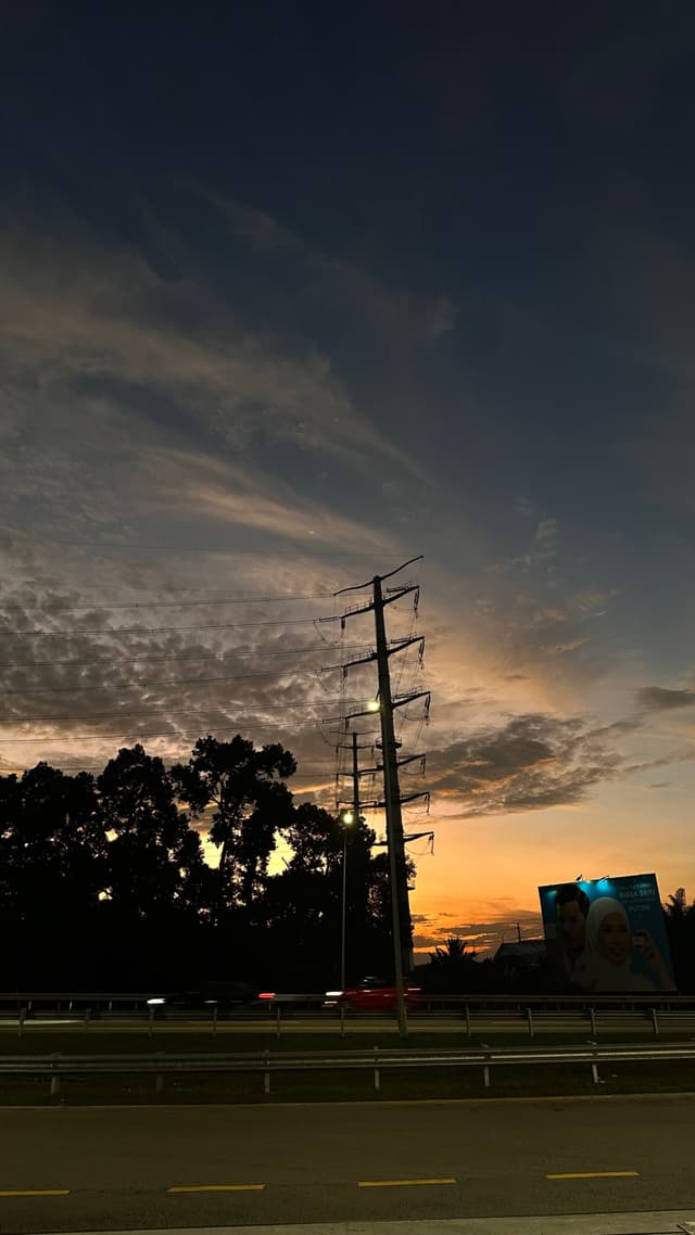 A power line standing tall on the side of a road under a sunset sky with clouds