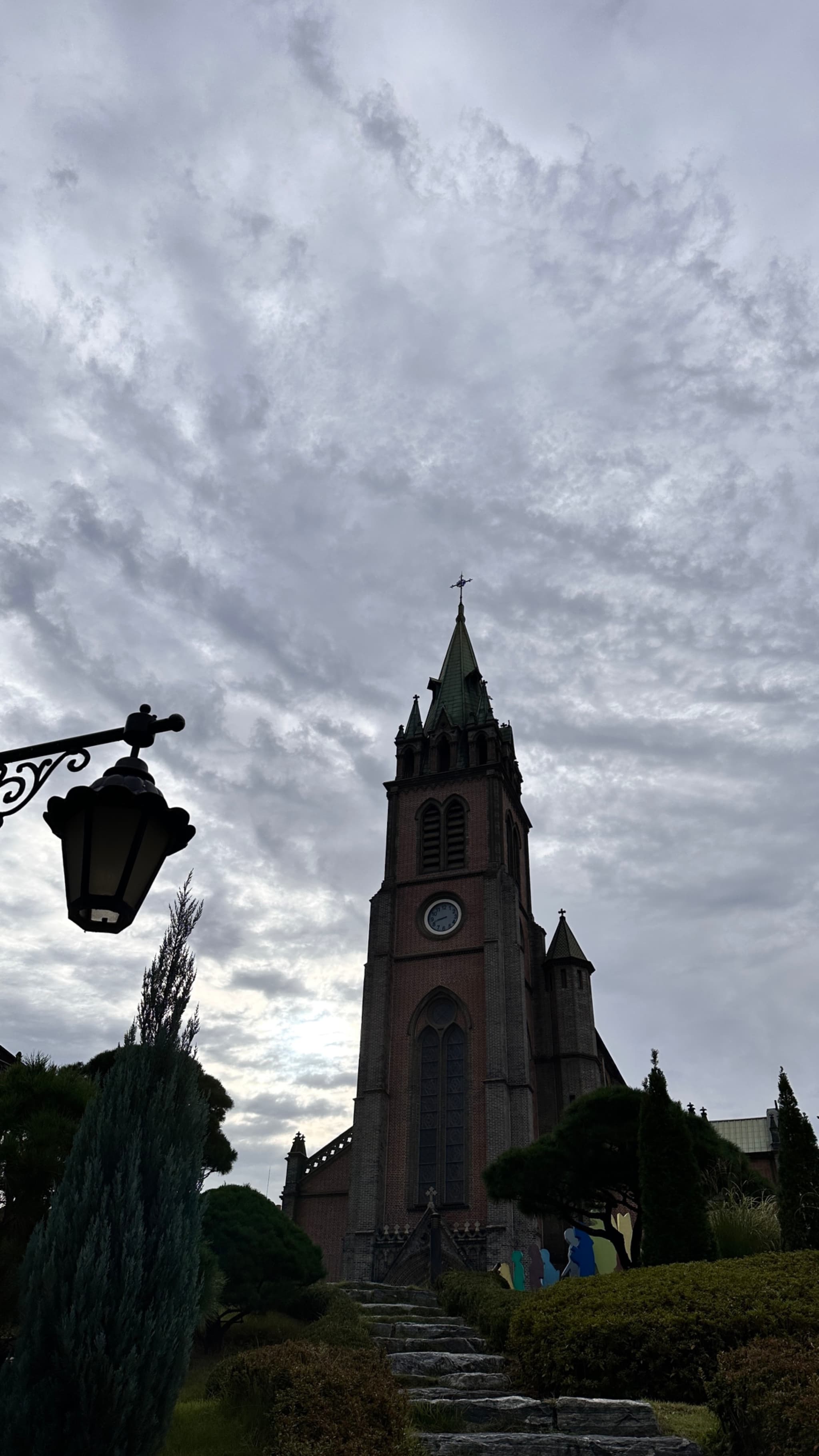 A church with a tall tower and clock face stands at the end of a stone pathway, surrounded by manicured shrubs and trees under a cloudy sky. A lamp post is visible on the left side