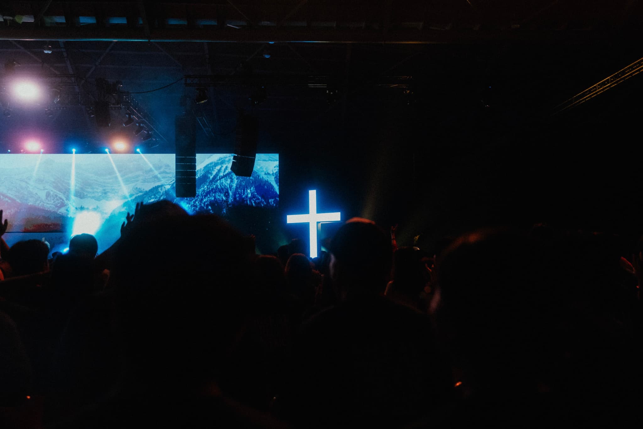 A dimly lit crowd with a glowing blue cross in the center and a lit-up screen in the background