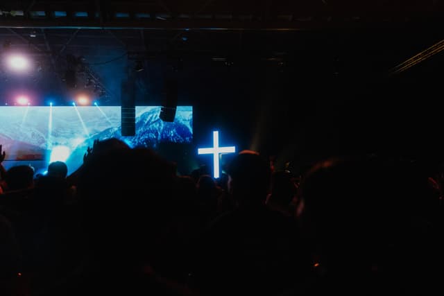 A dimly lit crowd with a glowing blue cross in the center and a lit-up screen in the background