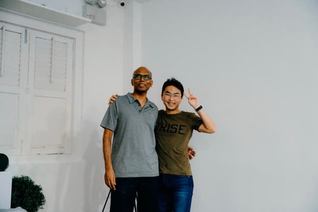 Two men standing side by side, one with his arm around the other's shoulder and the other with a hand raised in a wave, in front of a white wall and door