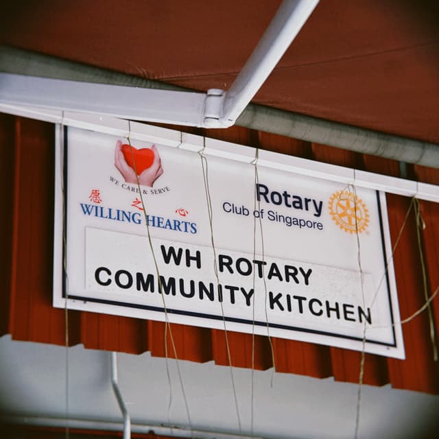 A white sign with a black border hangs from a building, displaying text and logos. The sign reads WH ROTARY COMMUNITY KITCHEN in bold black letters. Above this, two logos are visible: one features a red heart with hands cradling it, accompanied by the words WILLING HEARTS; the other displays an orange wheel alongside the text Rotary Club of Singapore. The sign is attached to a white gutter and appears to be situated outside a building with red slatted siding
