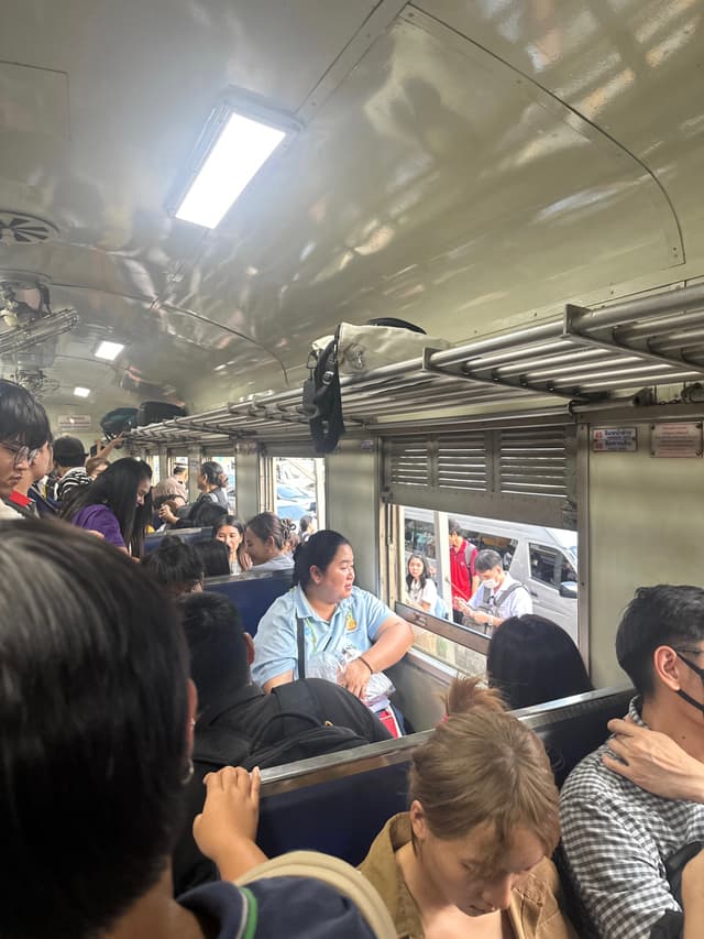 The interior of a crowded train car with people seated on either side. A man sits by a window looking at a book. The train has metal walls and a ceiling with fluorescent lighting