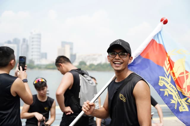 A smiling individual wearing a black cap and sleeveless top is holding a flagpole with a colorful flag. In the background, there are several people and a cityscape near a body of water. One person appears to be taking a photograph with a smartphone