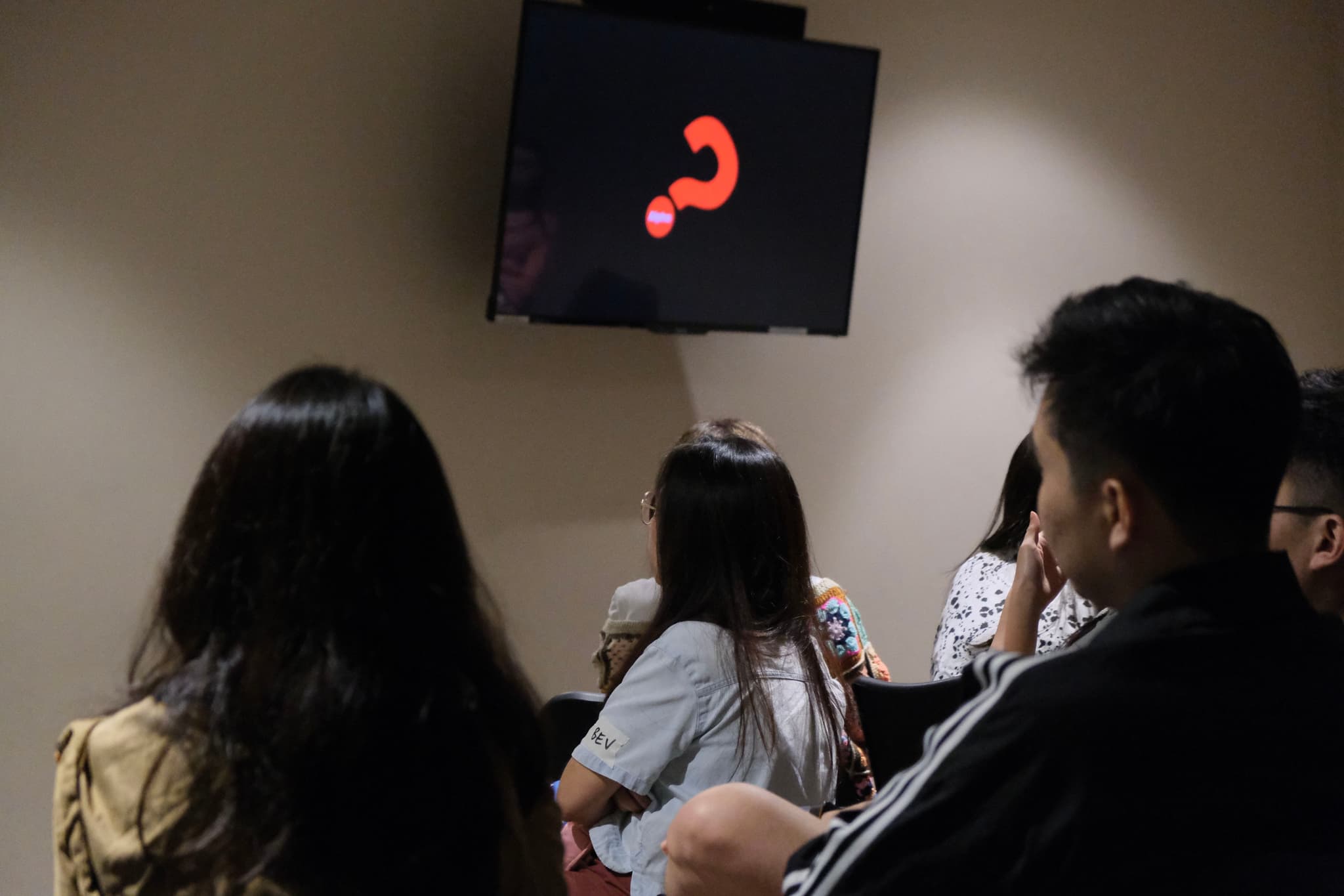Several individuals seated in a dimly lit room are facing a monitor displaying a large red question mark