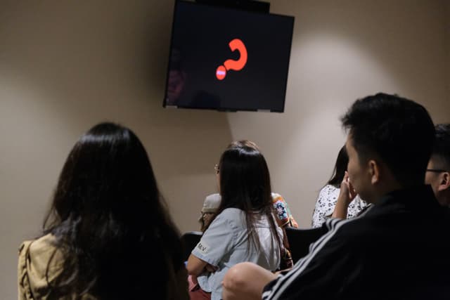 Several individuals seated in a dimly lit room are facing a monitor displaying a large red question mark