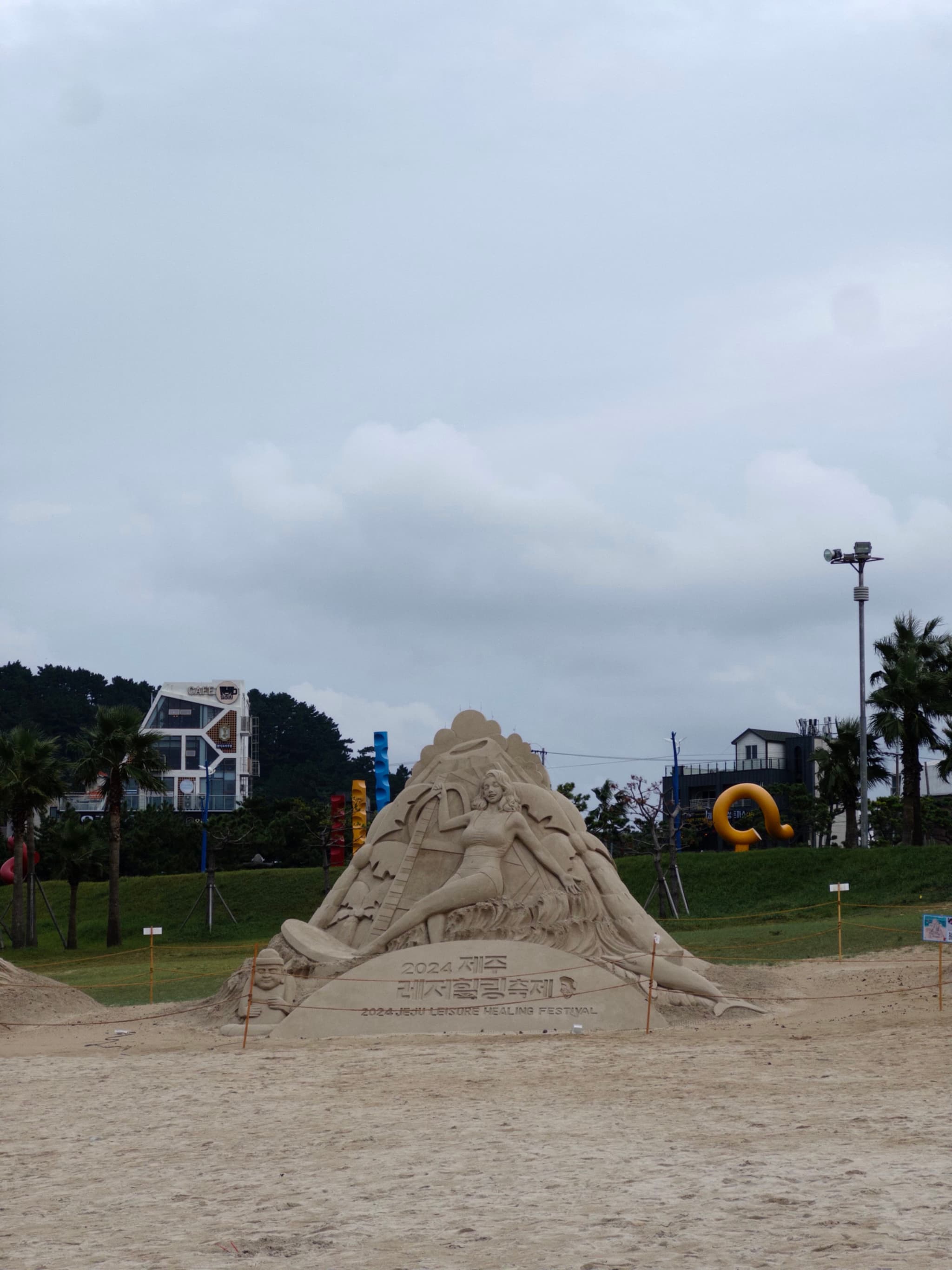 A large sand sculpture of some kind sits on a sandy beach with buildings and greenery in the background under a cloudy sky