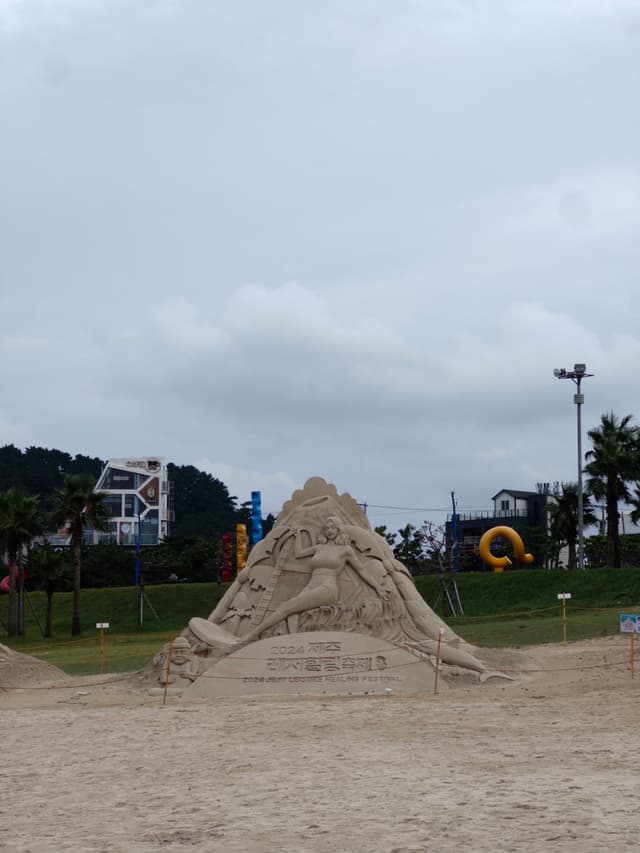 A large sand sculpture of some kind sits on a sandy beach with buildings and greenery in the background under a cloudy sky