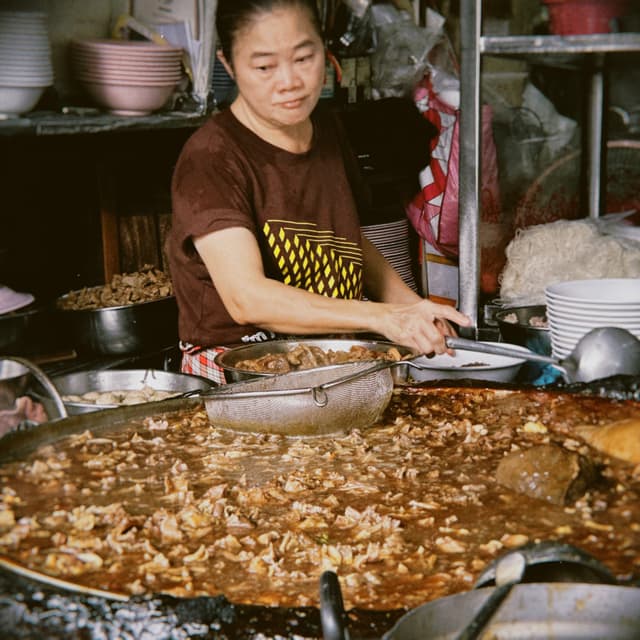 A woman mixes a large quantity of food in a wok. She wears a brown T-shirt and a yellow and black apron. The food appears to be meat and vegetables in a brown sauce. Various kitchen utensils and supplies are visible in the background