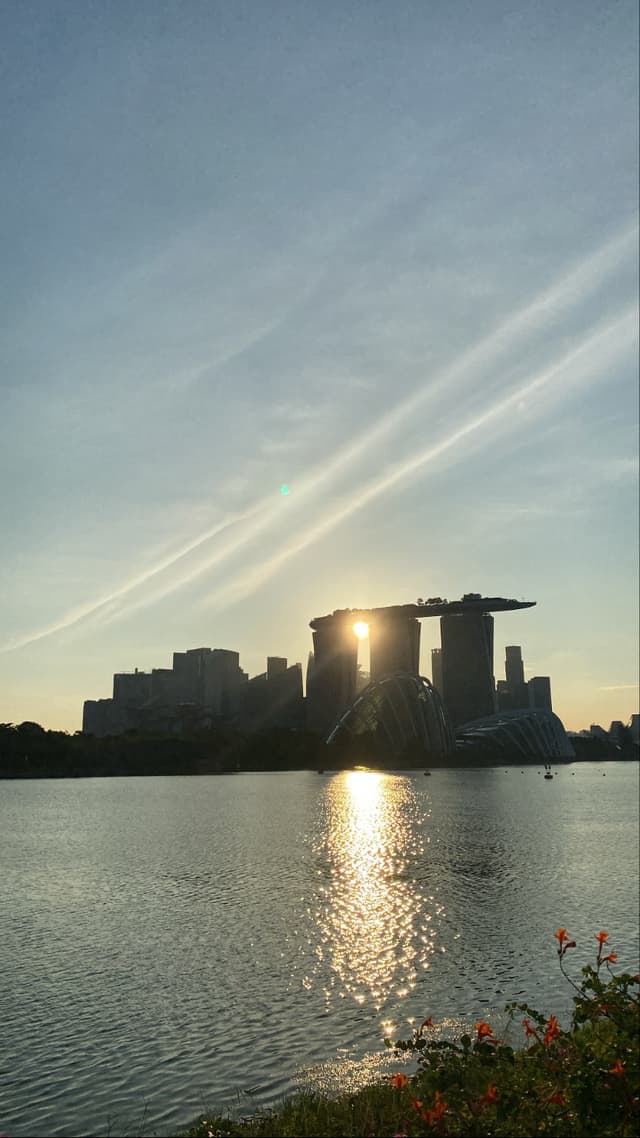 A sunrise or sunset over a calm body of water with the silhouette of a distinctive building featuring three towers connected at the top by a horizontal structure, likely the Marina Bay Sands in Singapore, with sunbeams radiating through the sky and reflecting on the water's surface