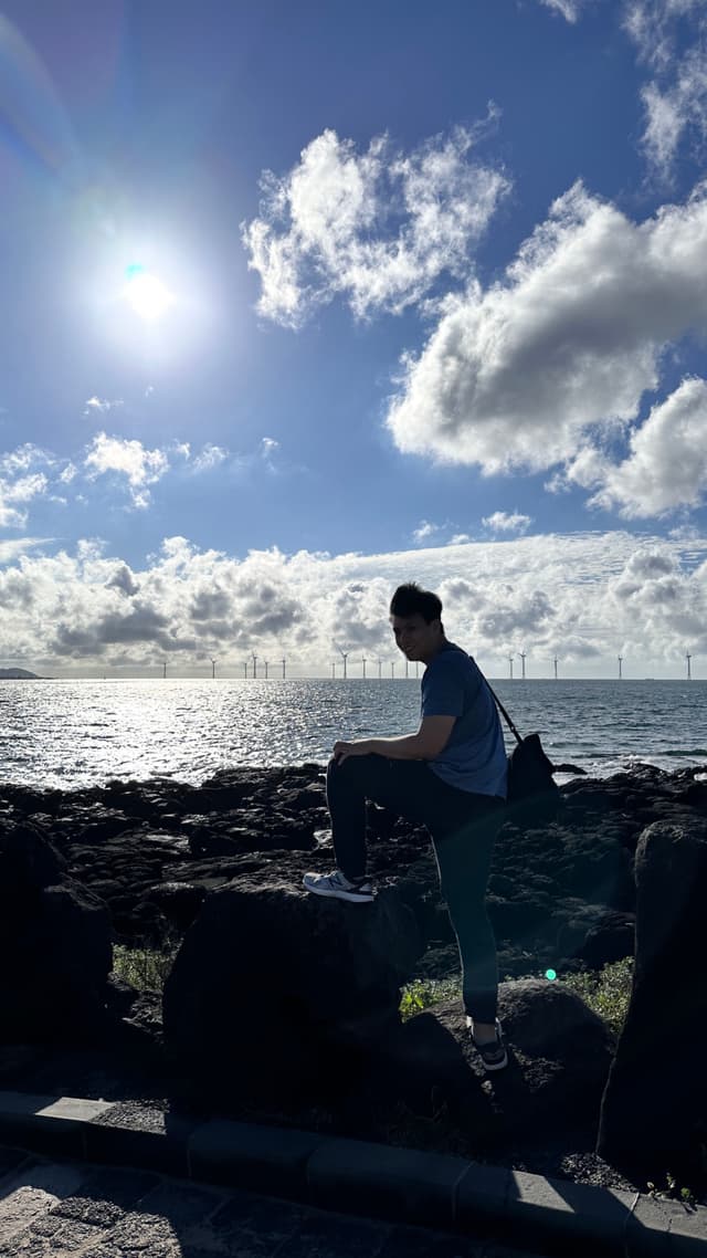 A man sits on a rock in front of a body of water, with a bridge visible in the background under a blue sky with clouds