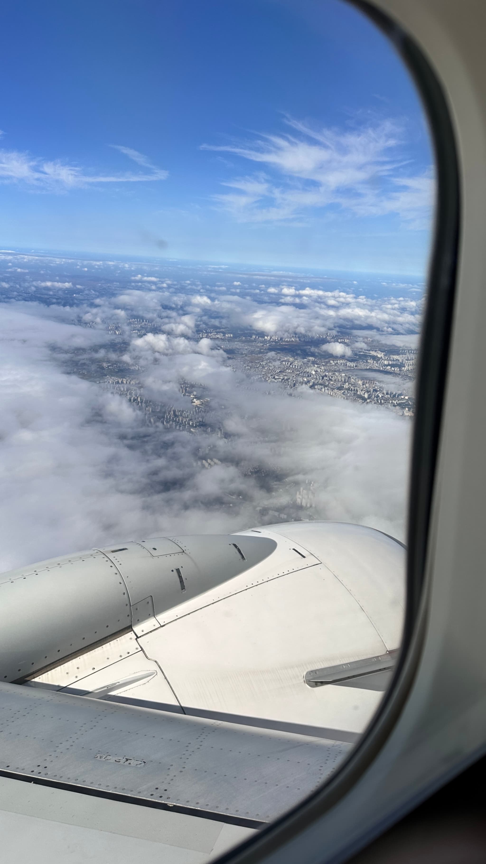 An airplane wing and engine are visible through a window, with clouds and a blue sky in the background. The wing and engine are white, with a view of the clouds and blue sky above. The image appears to have been taken from inside the plane