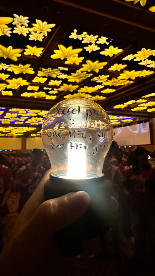 A hand holds a clear light bulb with a black base. The light bulb, engraved with text, glows brightly while being held up by a thumb and index finger. The background features a dark room with a ceiling adorned with hundreds of yellow maple leaf-shaped decorations and a crowd holding glowing objects