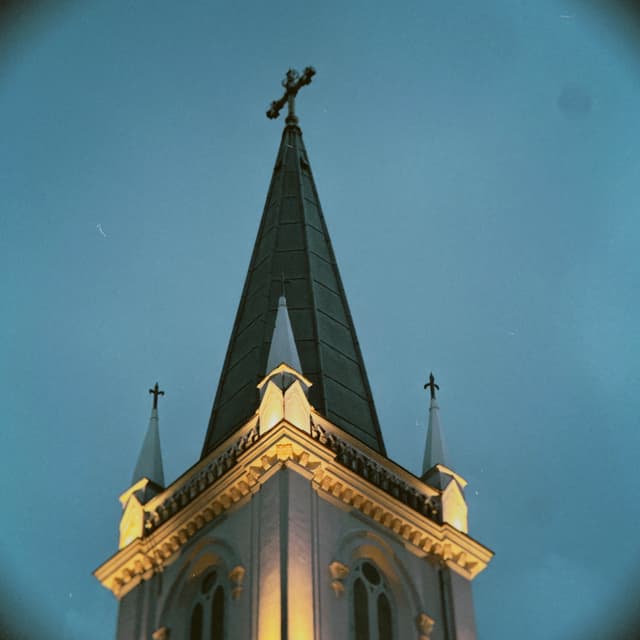 The top of a church with a tall, dark steeple and cross against a blue sky Smaller spires and crosses are visible on the sides The bottom of the building is lit up