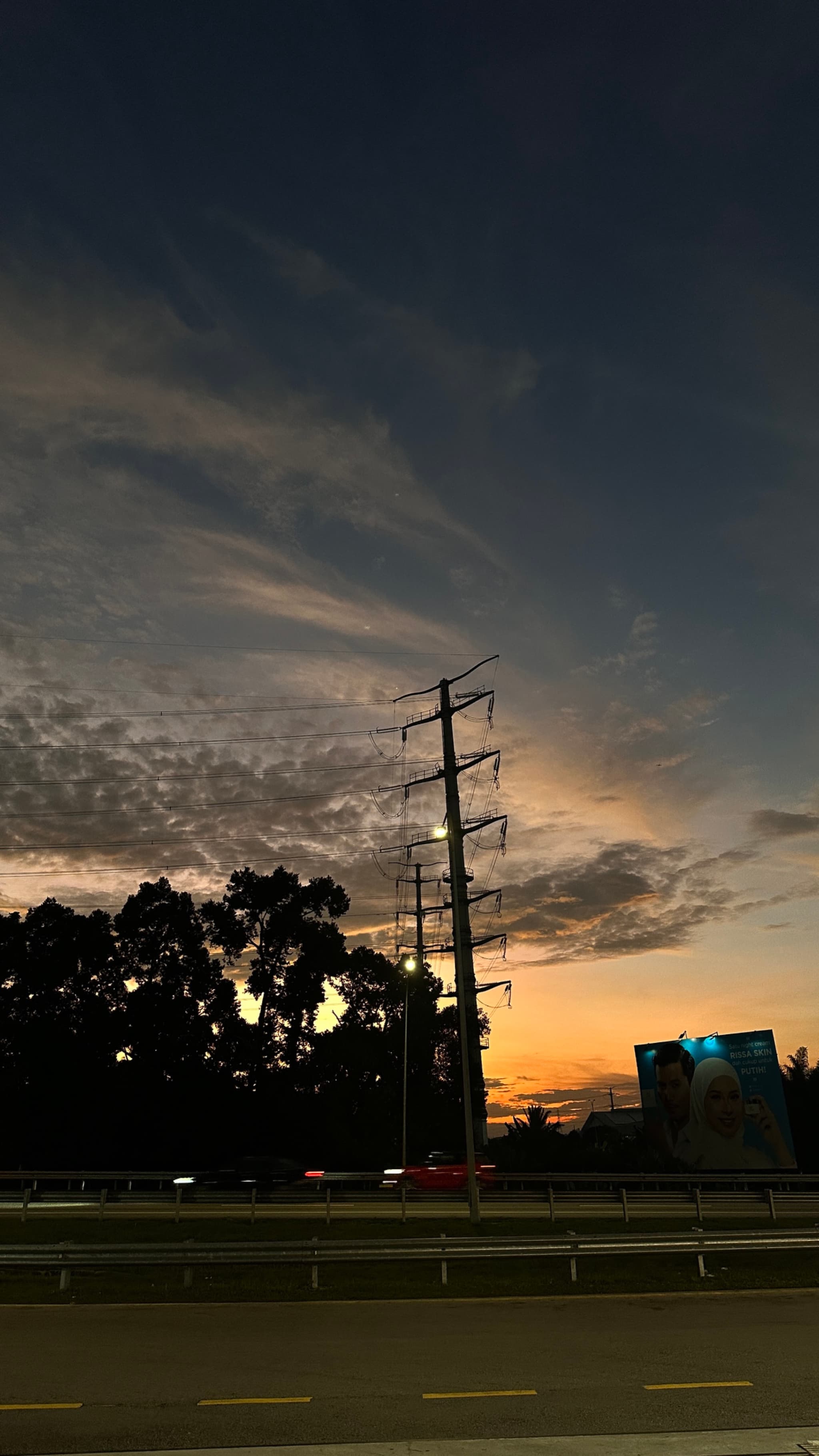 A power line standing tall on the side of a road under a sunset sky with clouds