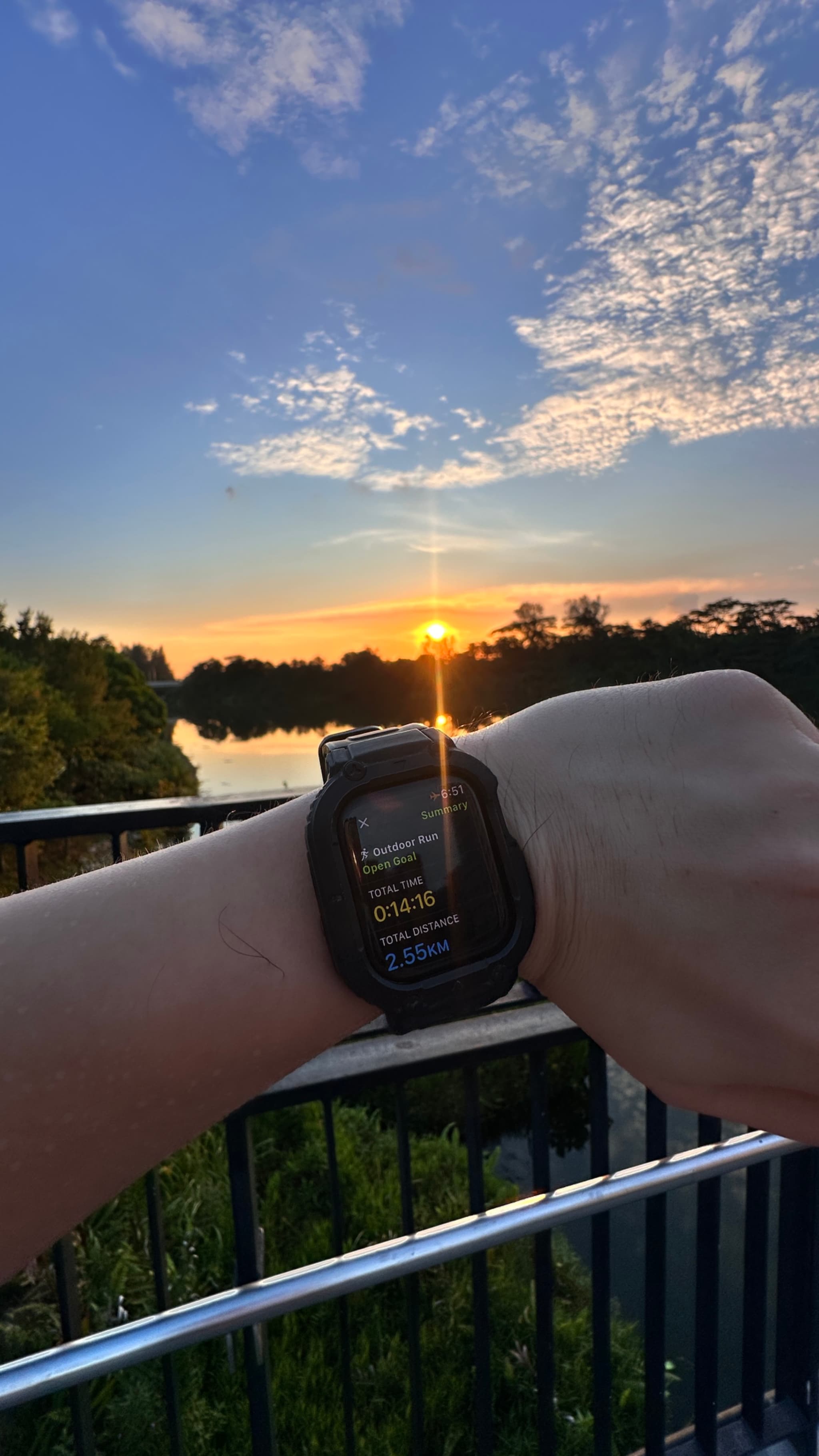 A person's arm with a smartwatch on their wrist, overlooking a serene lake or river at sunset The background features a blue sky with scattered clouds, and trees lining the water's edge A metal railing is visible in the foreground