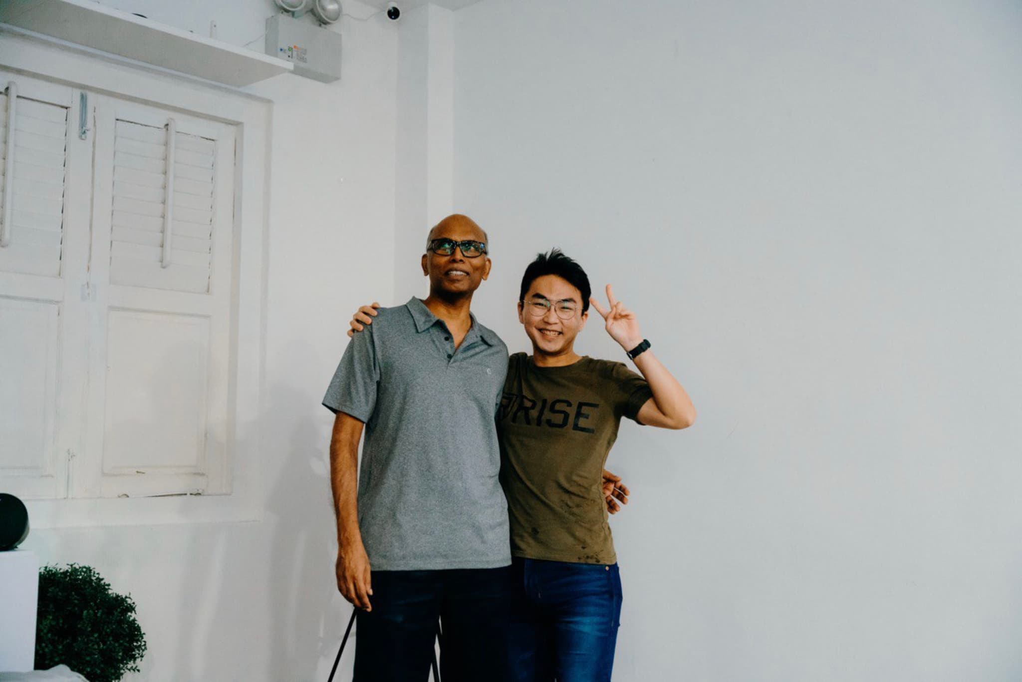 Two men standing side by side, one with his arm around the other's shoulder and the other with a hand raised in a wave, in front of a white wall and door