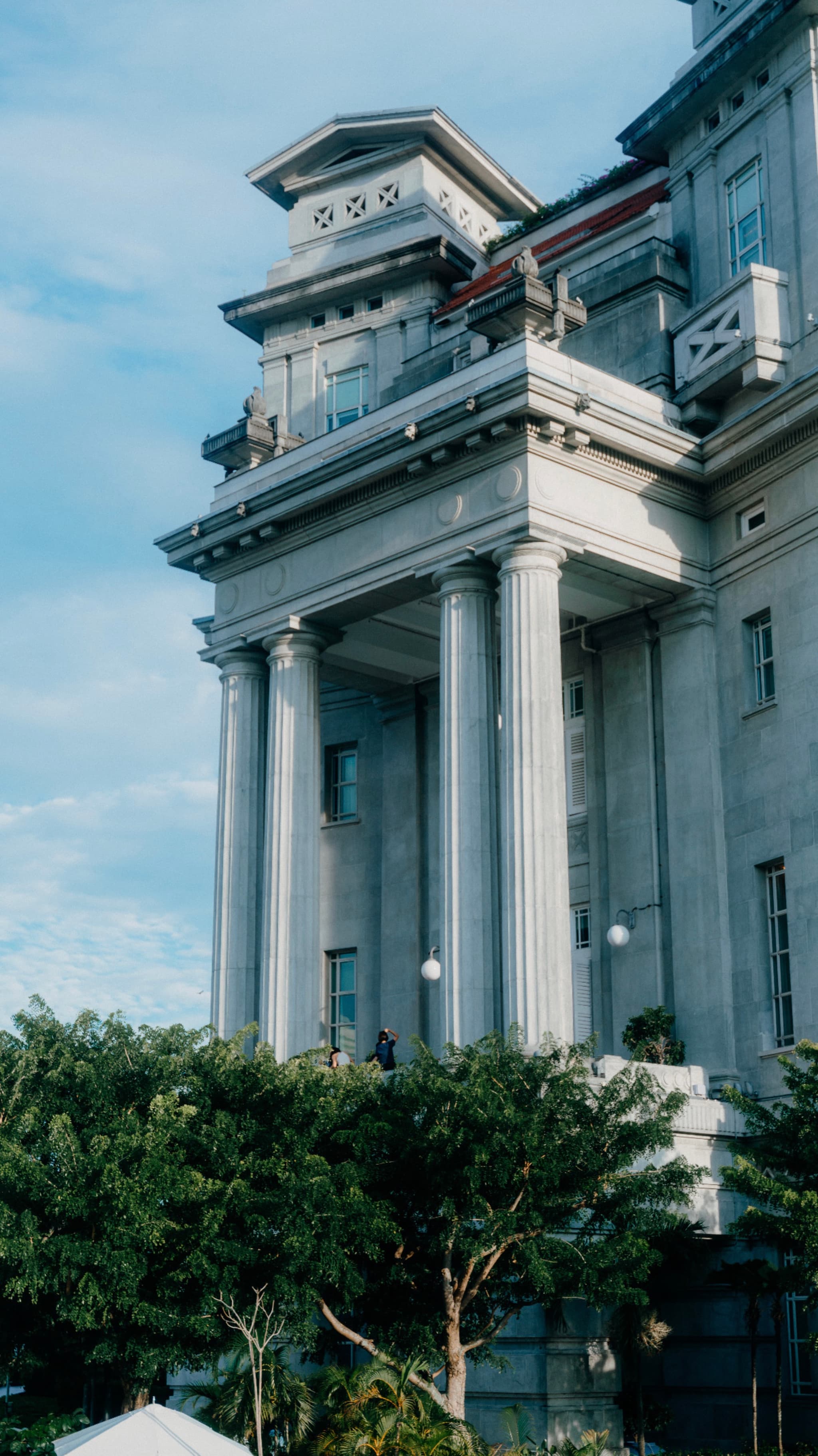 A large, gray building with columns and a tower on top, surrounded by trees and a blue sky