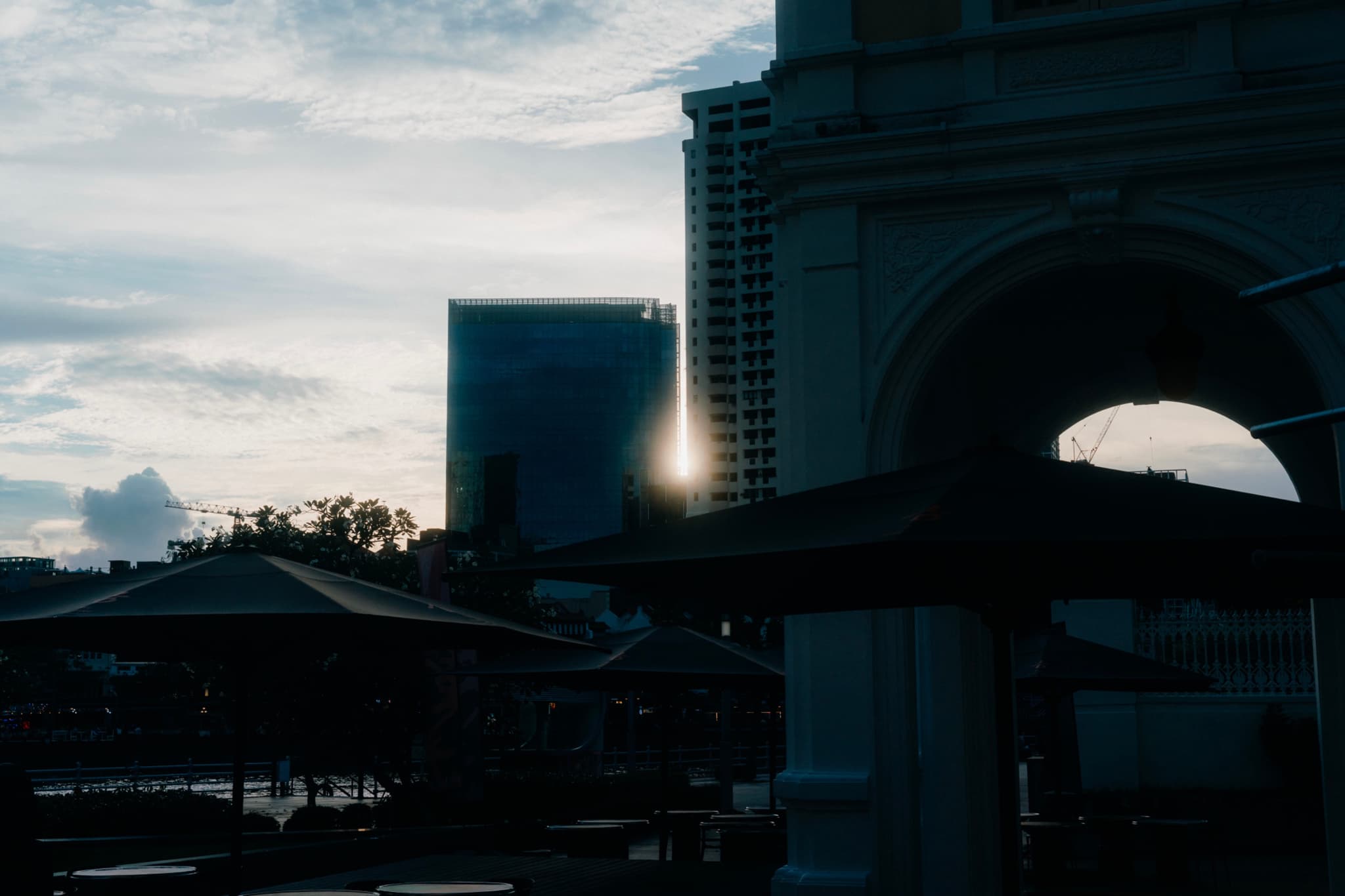 A darkened cityscape with a large building featuring an archway and a tall skyscraper in the background, set against a cloudy sky at sunset